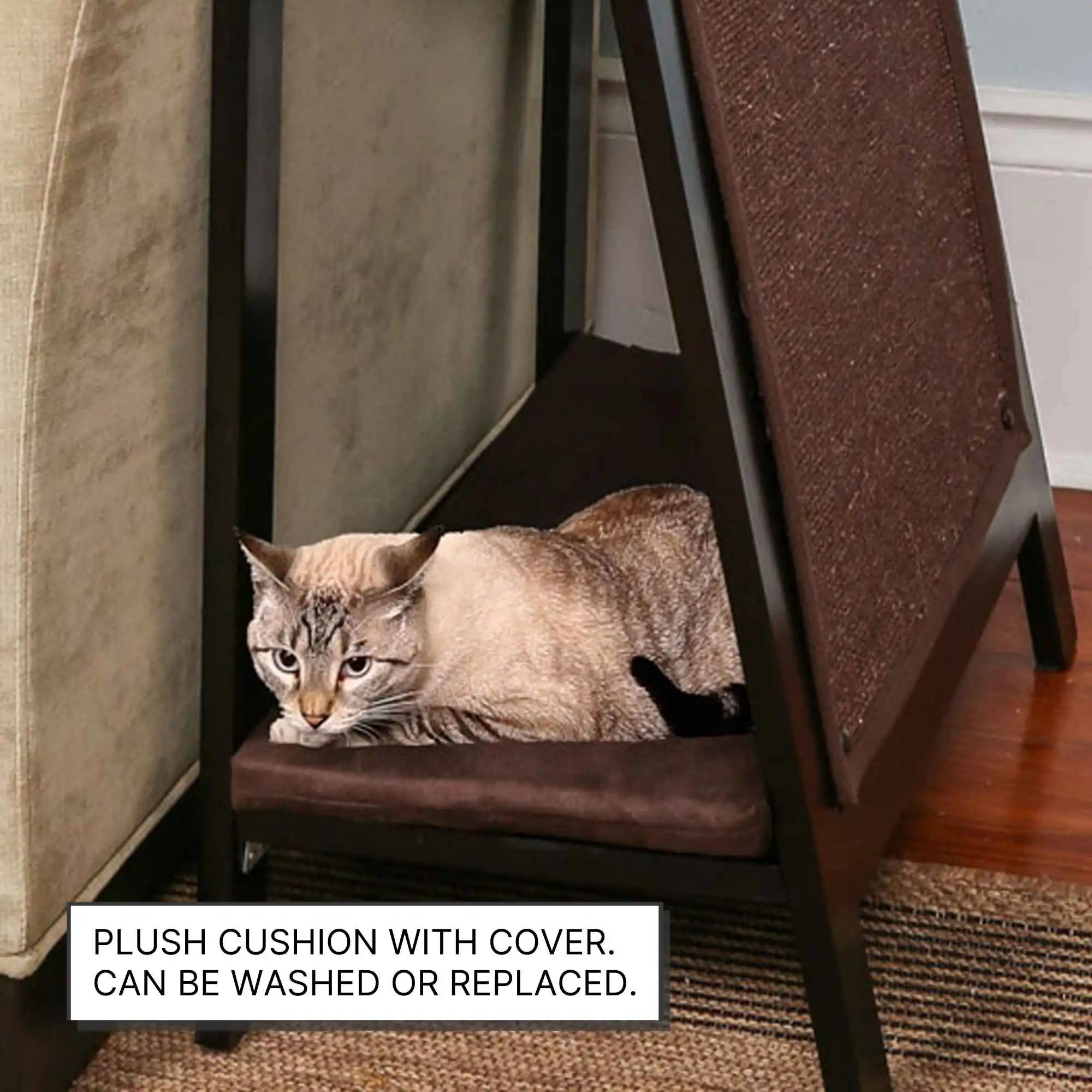 A cat relaxes on an A-frame wooden cat bed with a brown plush cushion under a table next to a beige couch.