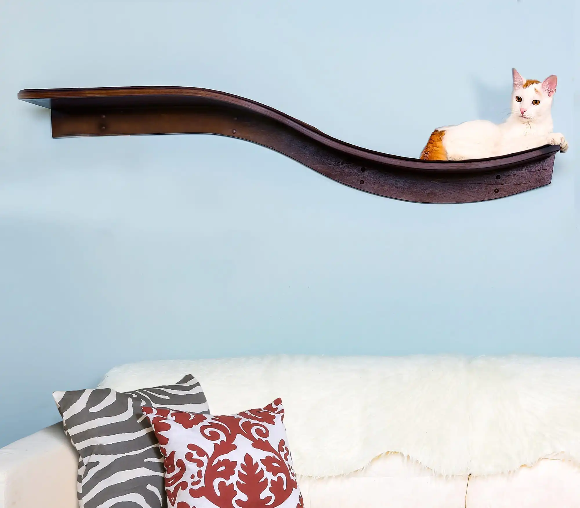White and orange cat lounging on a mahogany cat shelf mounted above a white sofa with patterned pillows and fur throw.