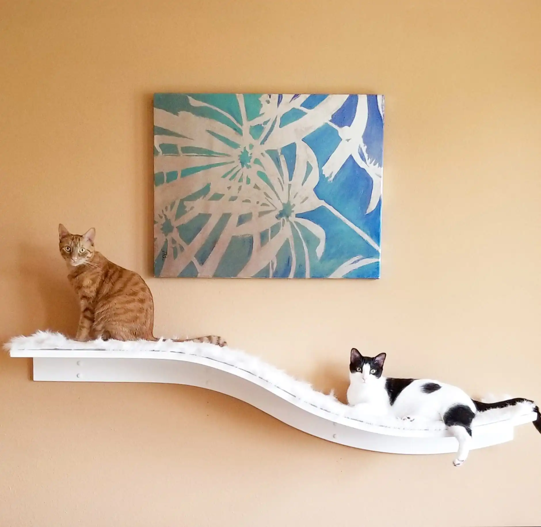 Two cats lounging on a white, fluffy Lotus Branch Cat Shelf beneath a blue abstract painting on a beige wall.
