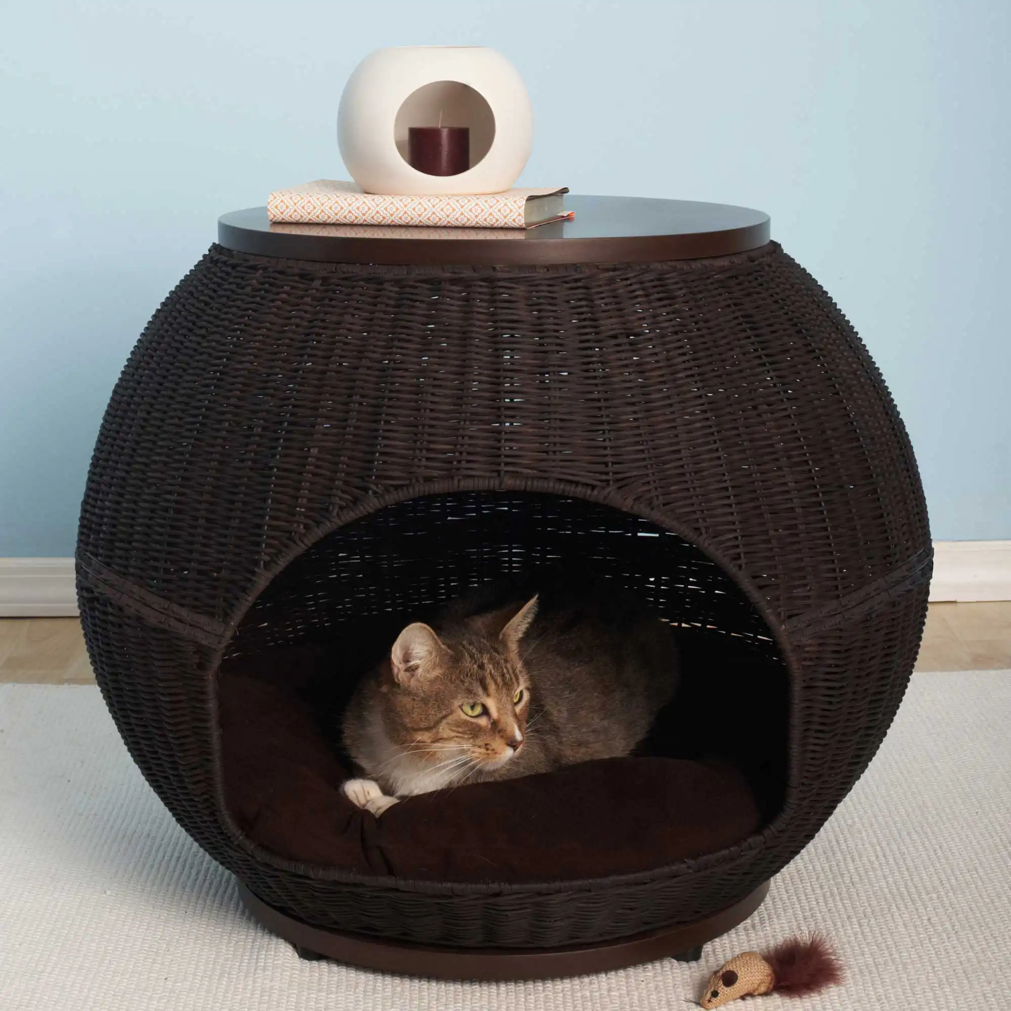 A tabby cat rests inside a round wicker bed that doubles as a side table holding a toy mouse, books, and a vase.