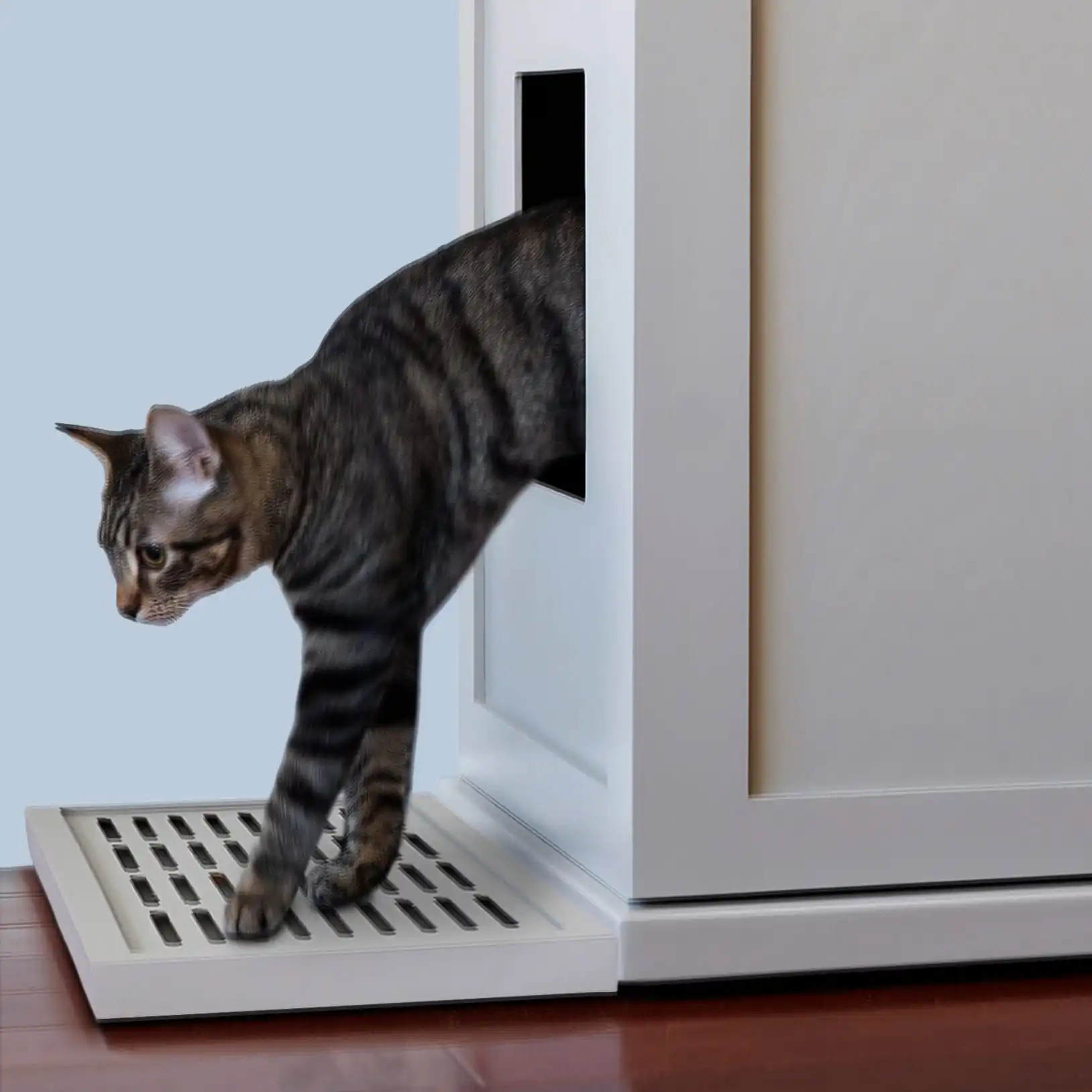 Tabby cat stepping from a white litter box cabinet onto a gray grated mat on a light wooden floor.