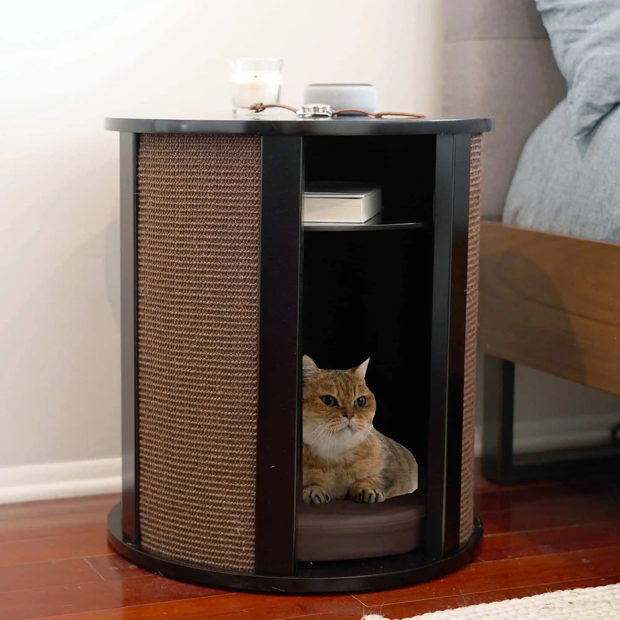 A cat lies inside a small round espresso side table with open shelves, next to a bed on a wooden floor.