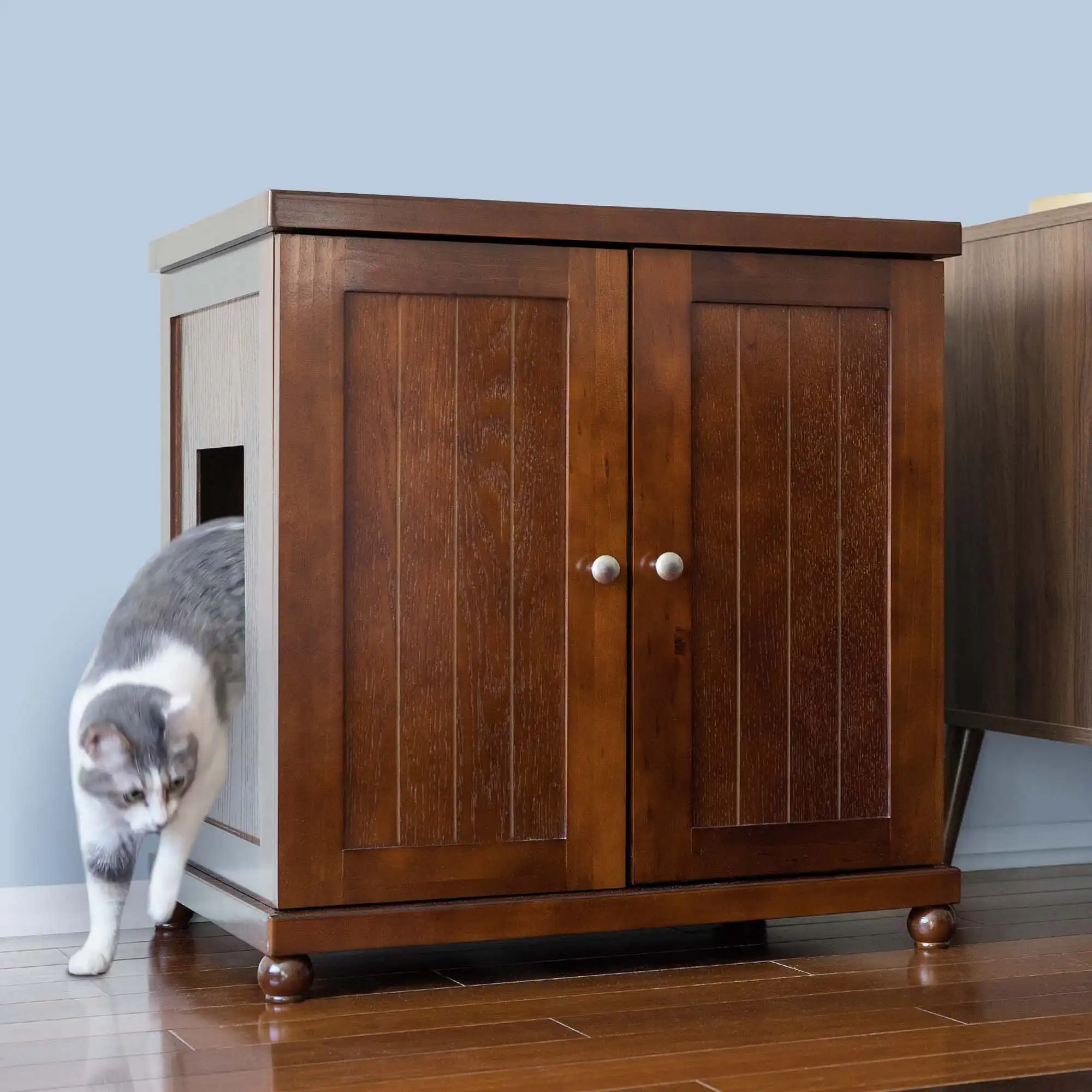 A gray and white cat steps out of a side opening in a wooden cabinet with double doors on a hardwood floor.