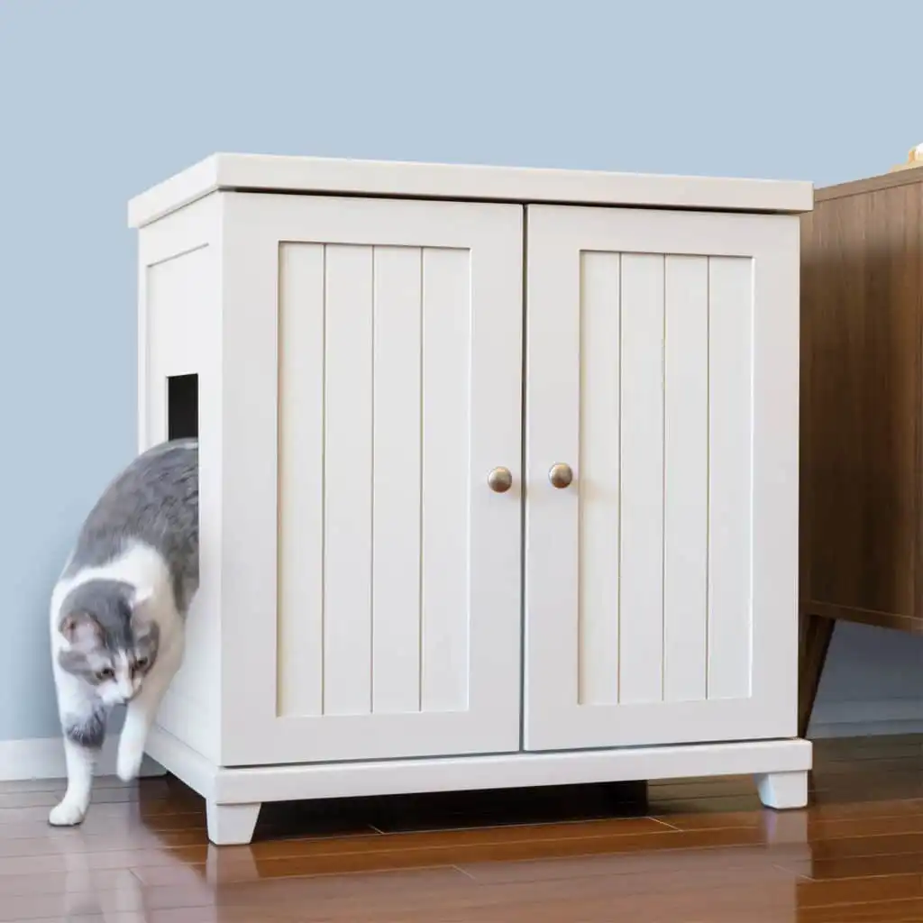 Gray and white cat exits side opening of sleek white two-door cabinet on wooden floor next to another furniture piece.