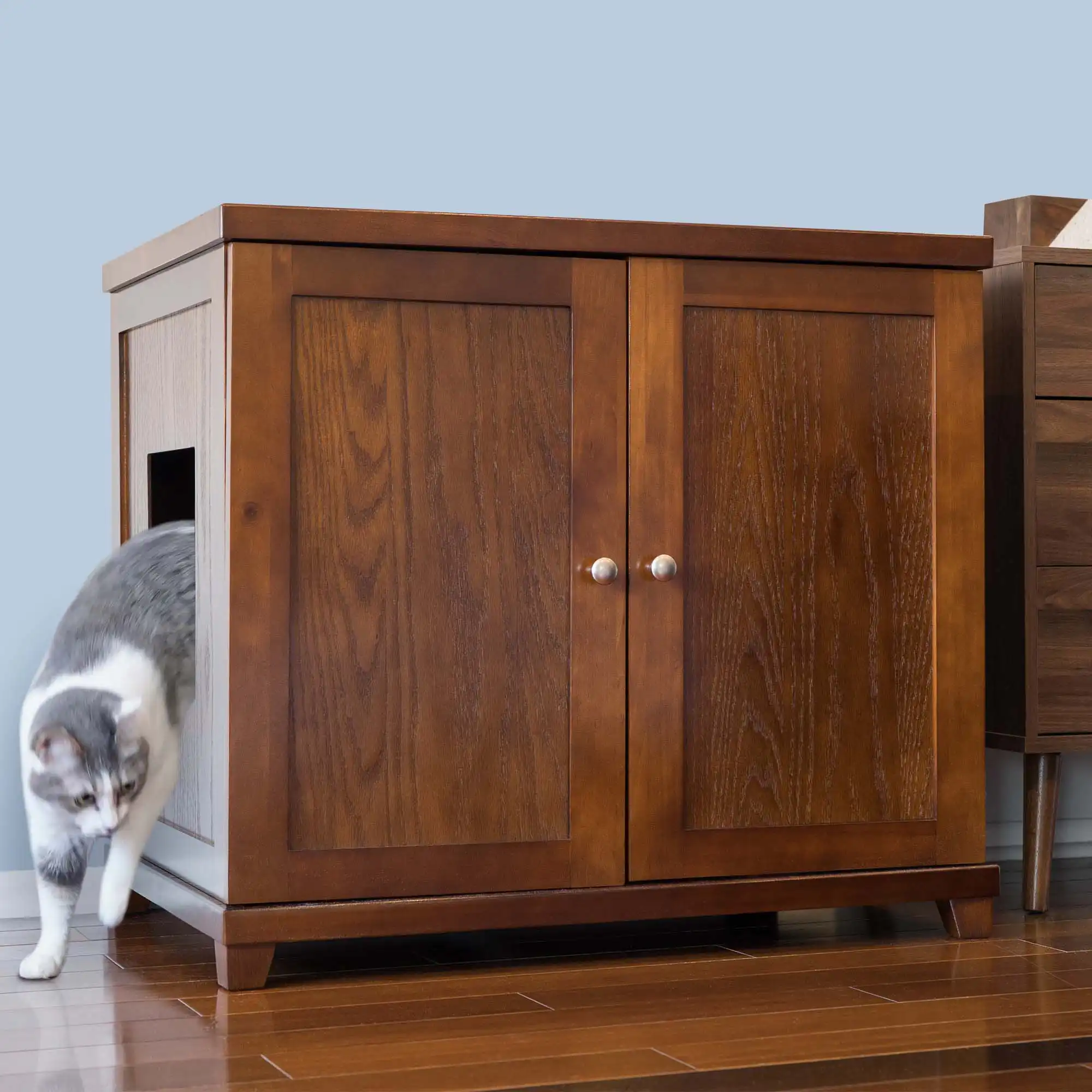 Gray and white cat walking out of a side opening in a wooden cabinet-style litter box on a hardwood floor.