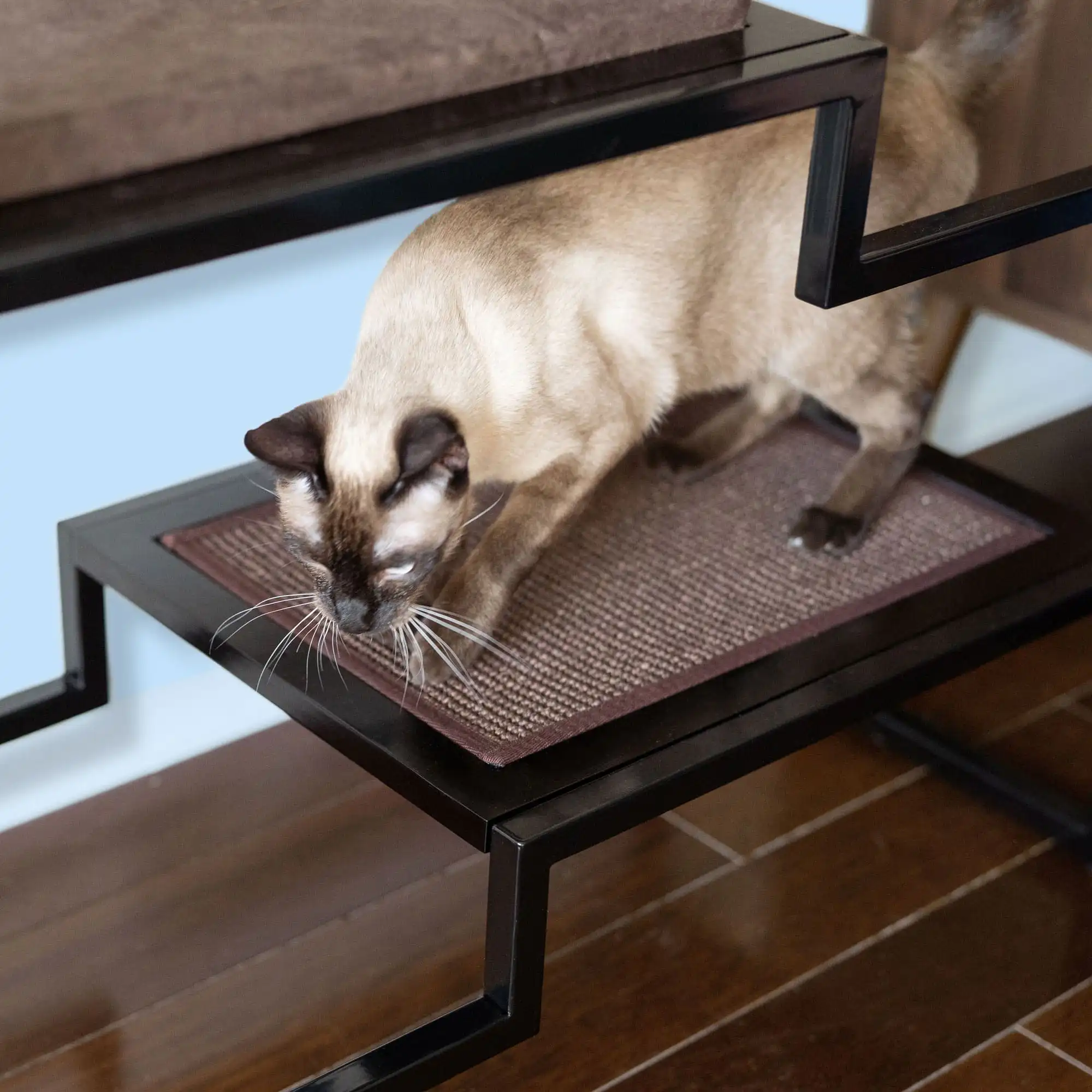 Siamese cat scratching a brown sisal mat on a black metal and wood cat condo set on a wooden floor.