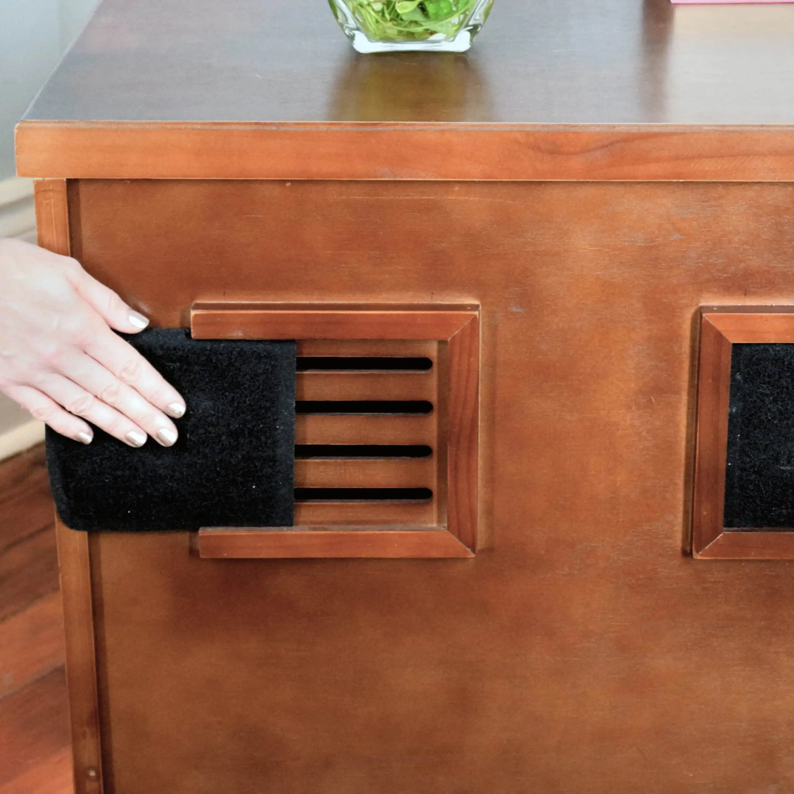 A hand inserts a Charcoal Litter Box Filter into a vent located on the side panel of a wooden cabinet.