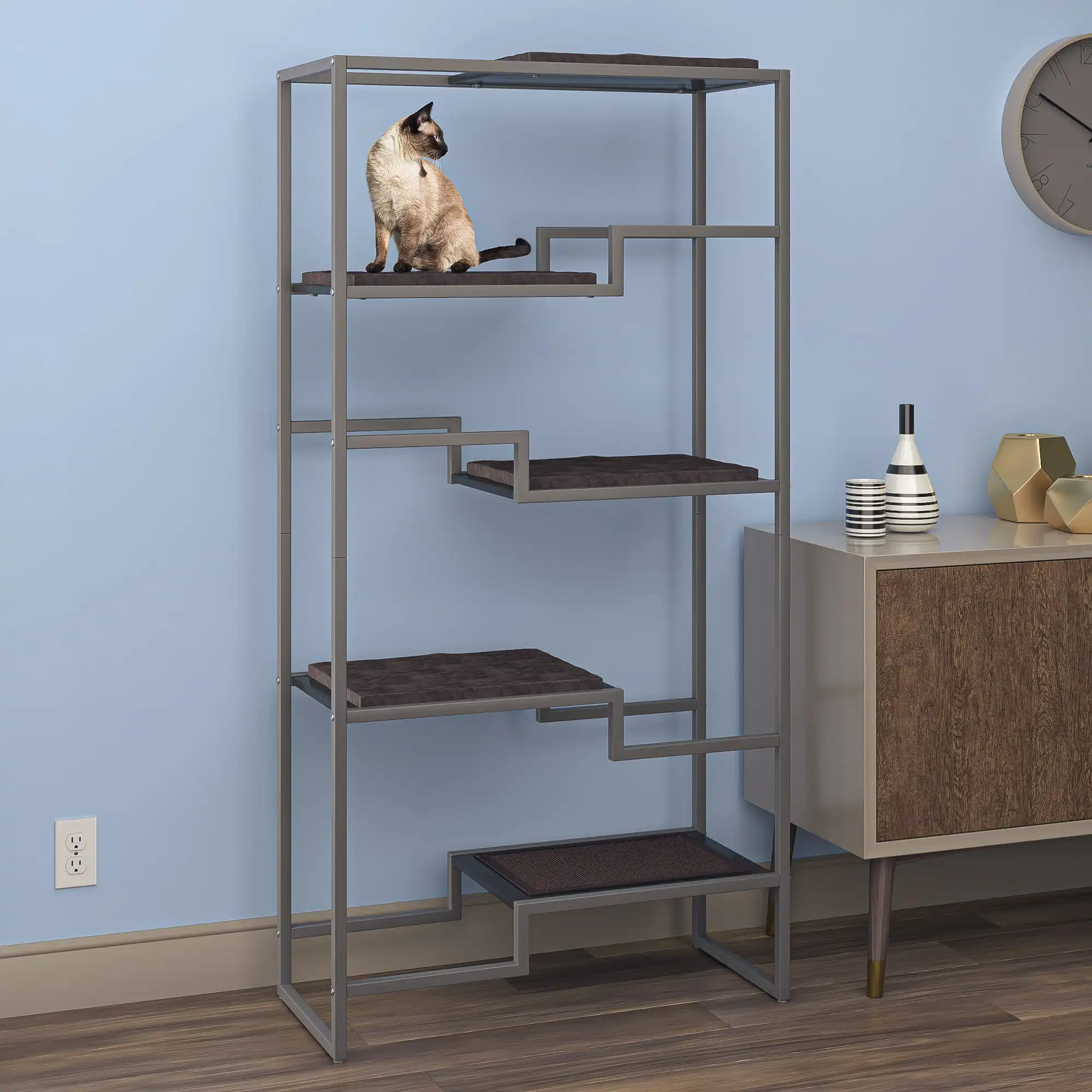 Siamese cat on top platform of gray metal cat tower with brown cushion, against a light blue wall in modern living room.