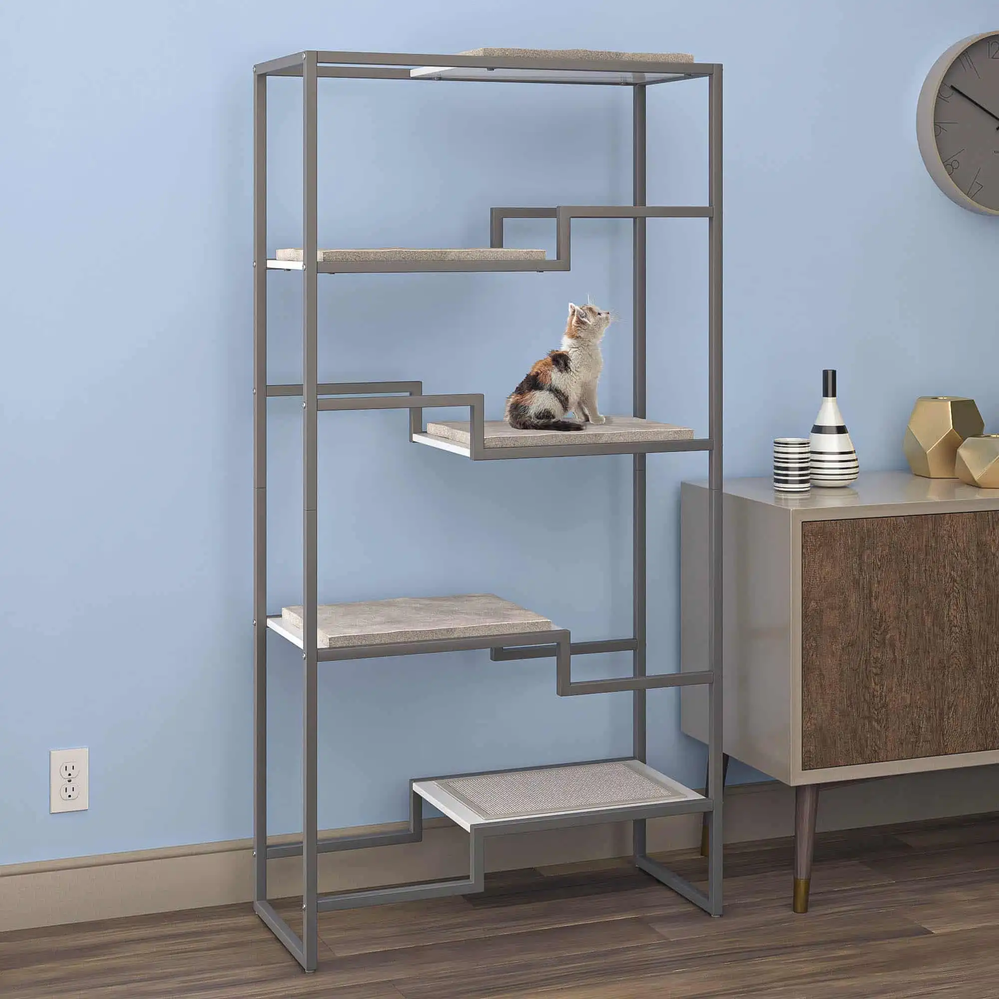 Cat sitting on a metal and wood cat condo with gray frame, white platform, gray cushion; near a cabinet and wall clock.