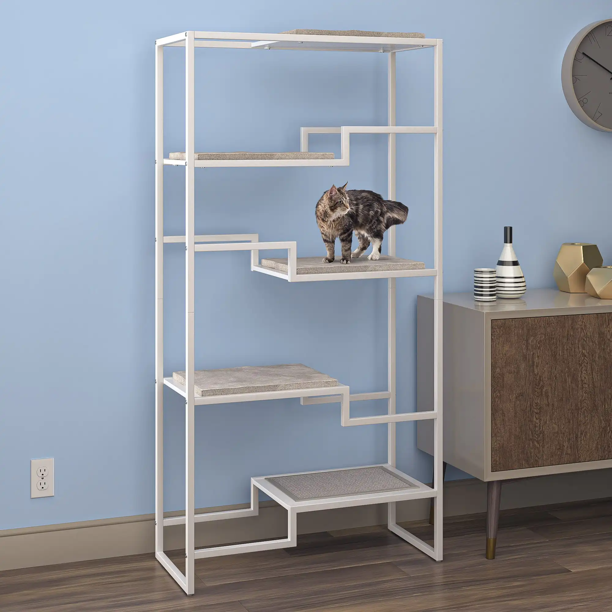 A cat stands on the second shelf of a white-framed, gray-cushioned condo near a wooden cabinet and blue wall.