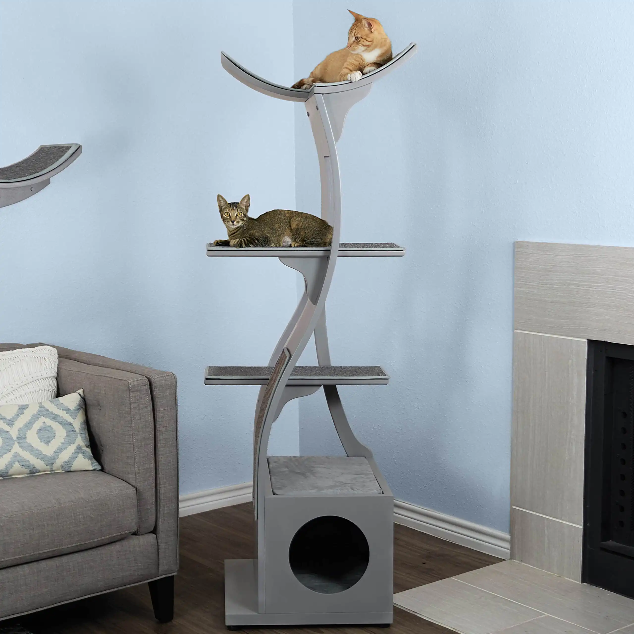 Two cats rest on separate tiers of a smoke gray cat tower between a sofa and fireplace in a light blue living room.