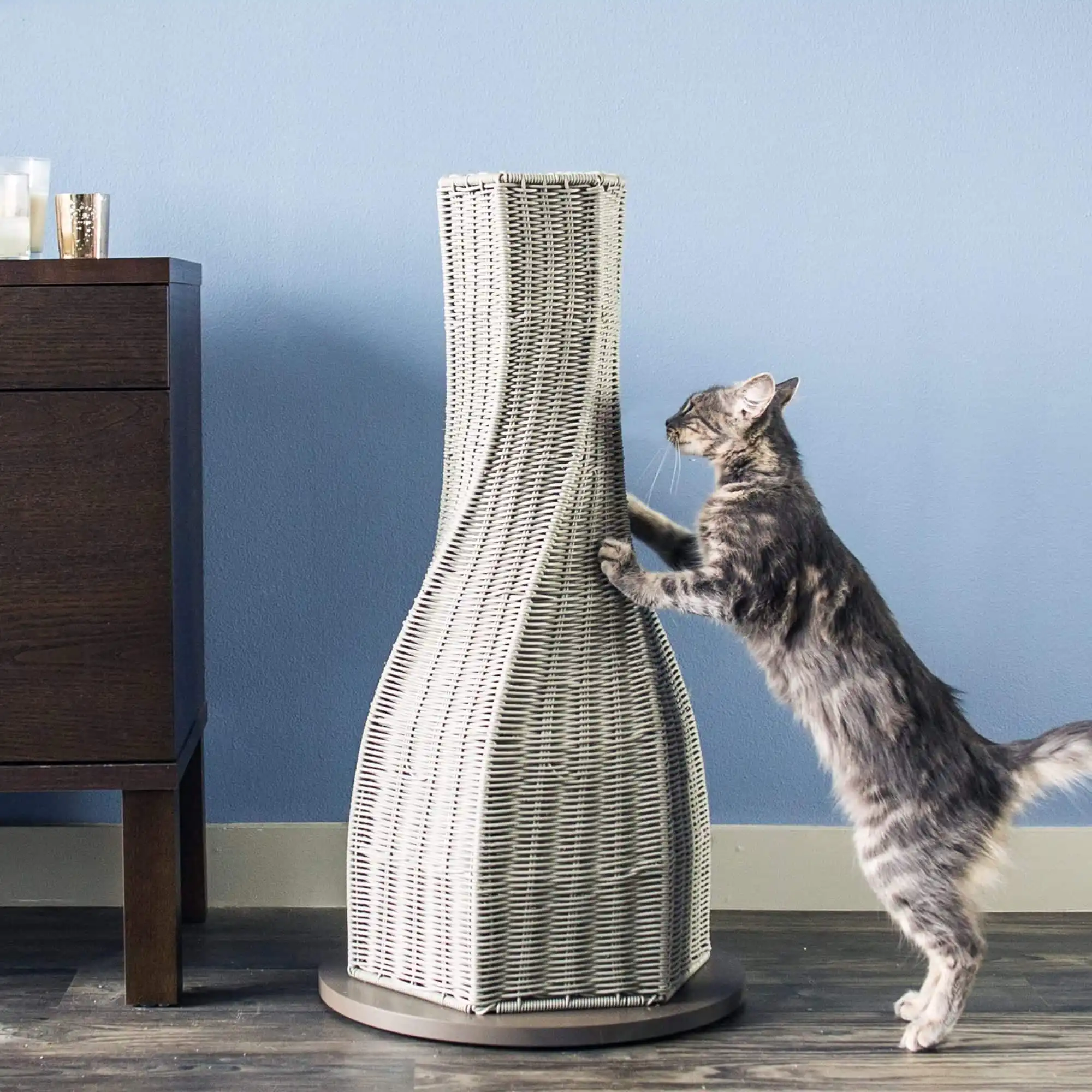 A cat stretches on a tall, woven scratching post next to a dark wooden cabinet against a blue wall.