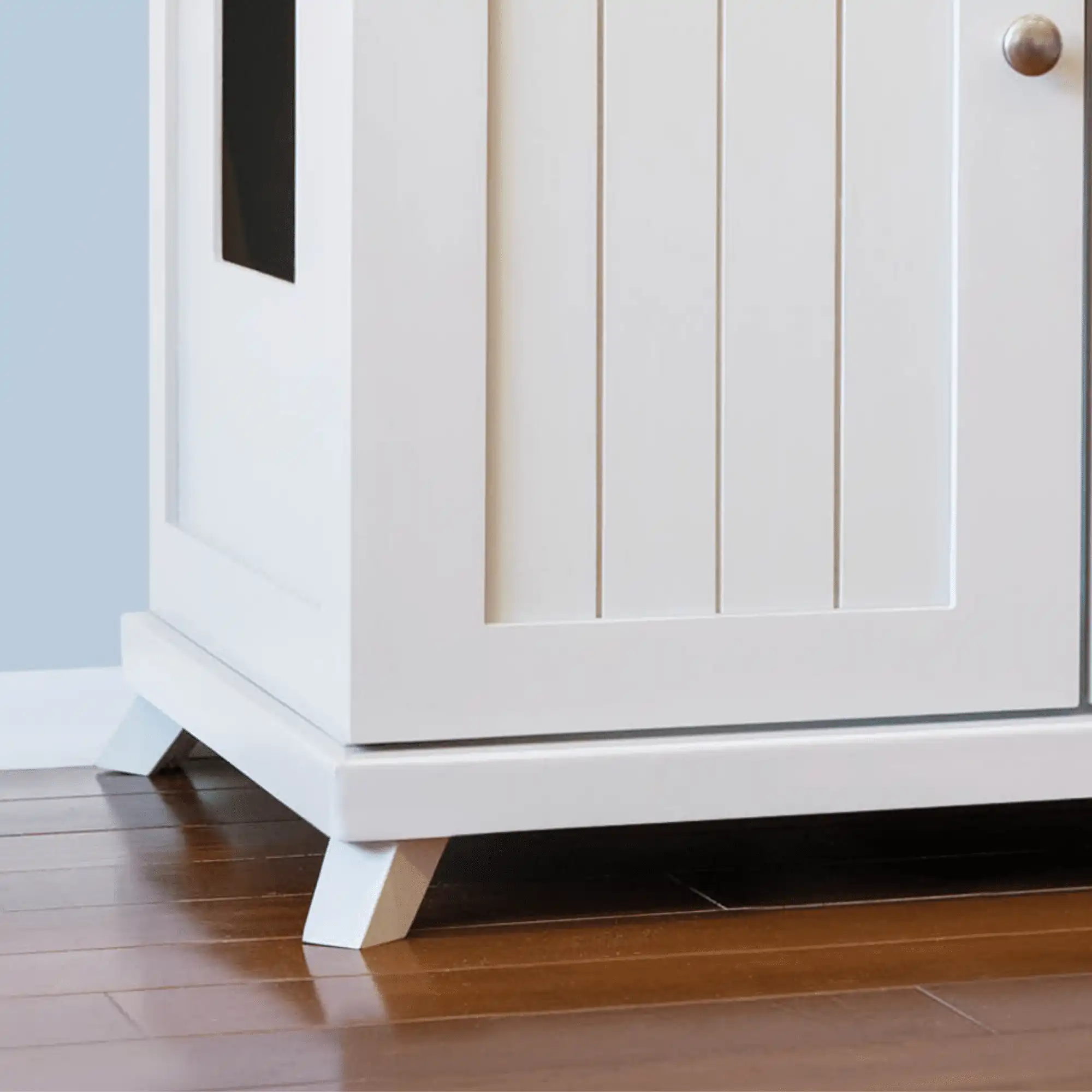 White wooden cabinet with paneled doors and four short angled legs on a polished hardwood floor.