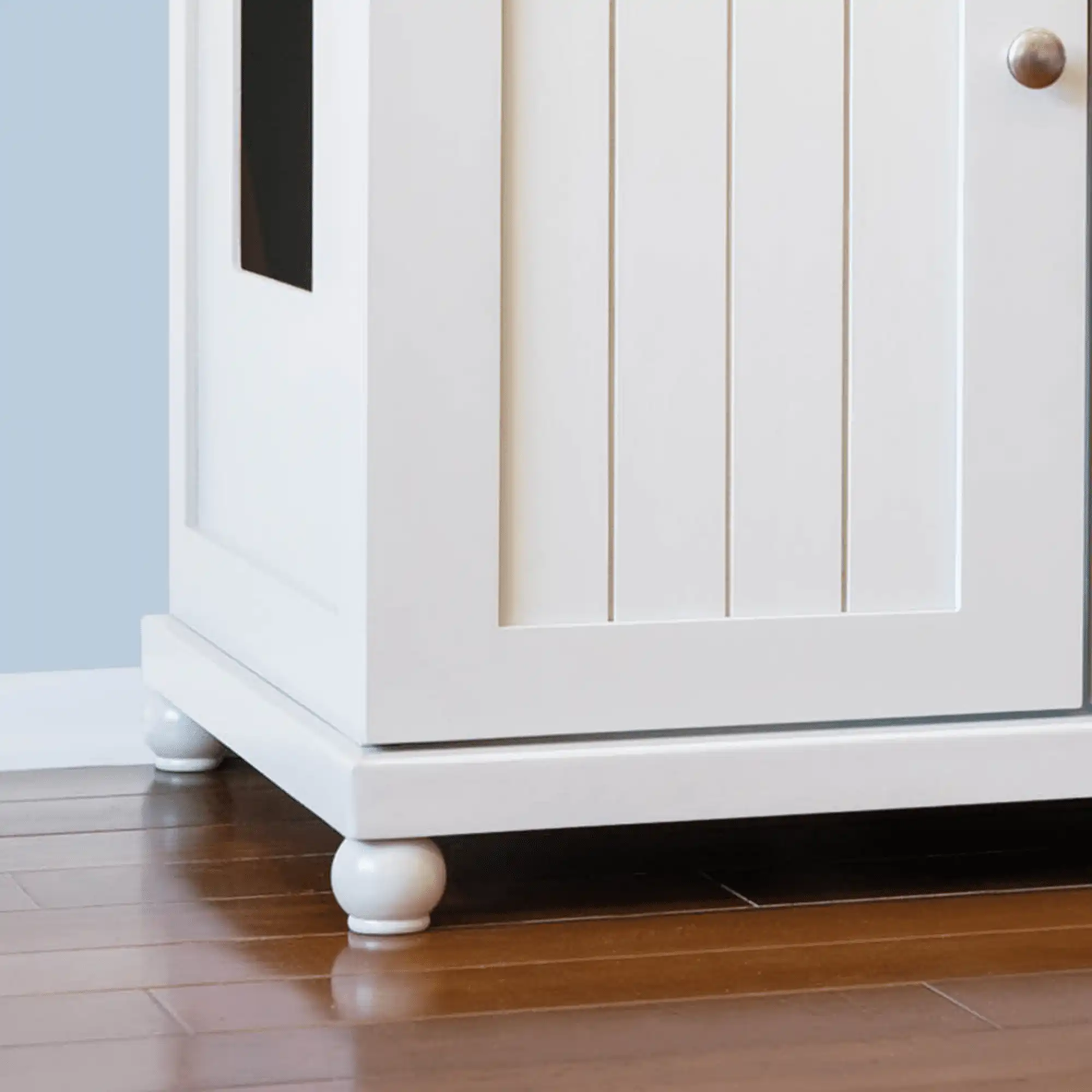 Deluxe white cabinet with panel door, round knob and feet, side rectangular opening, on a hardwood floor.