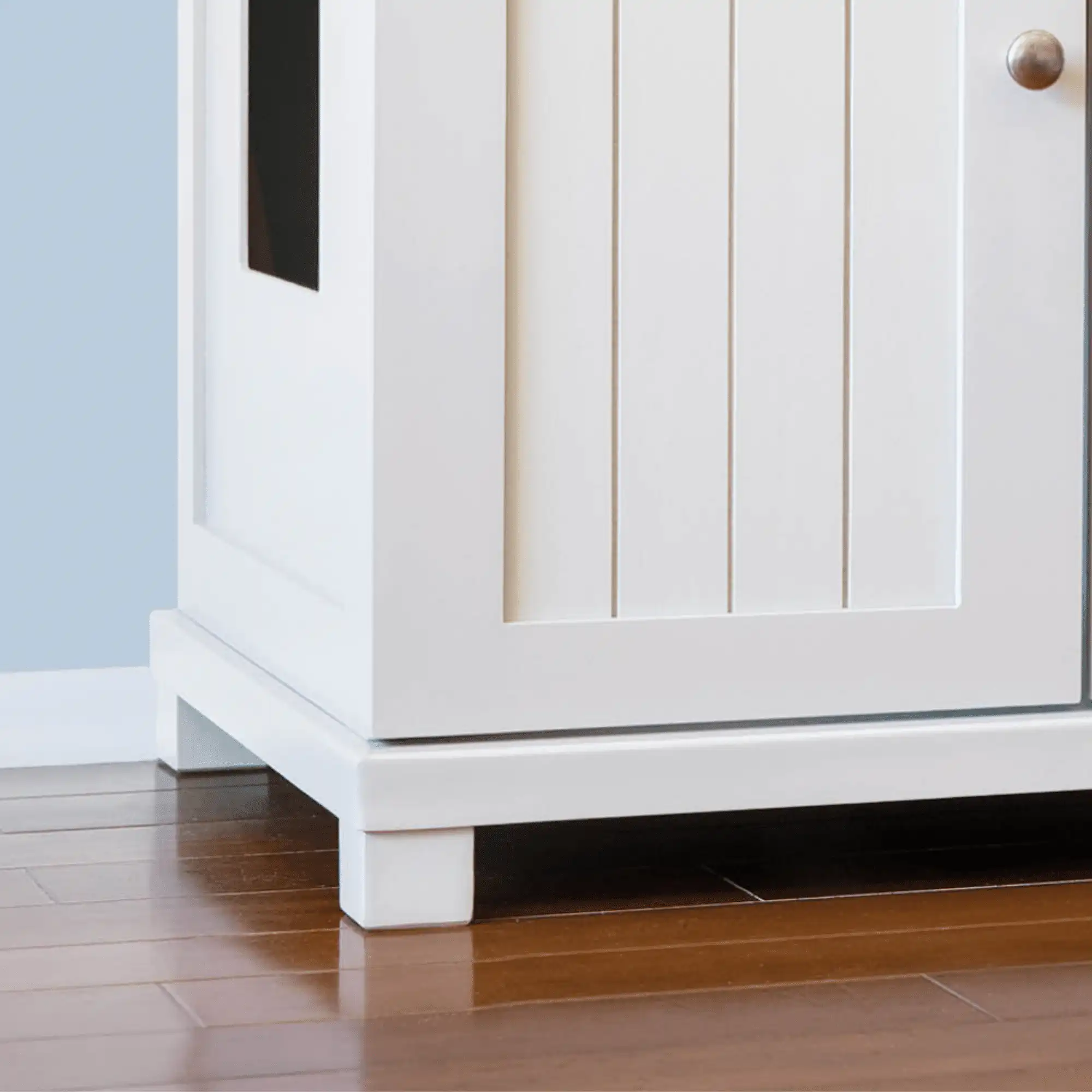 White wooden cabinet with feet, vertical door, round handle, side cutout on brown floor against light blue wall.