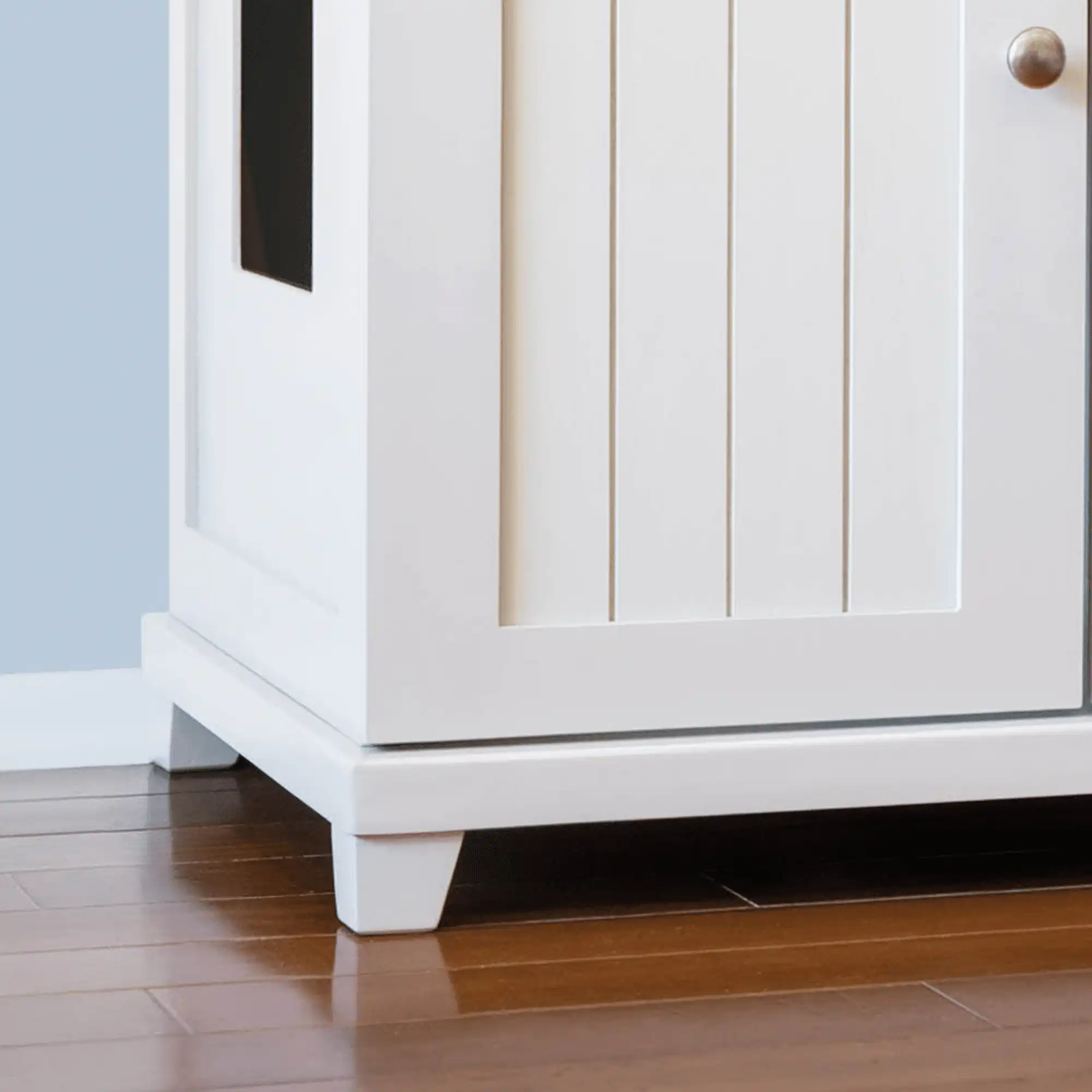 White cabinet with vertical groove doors, metal handle, cat litter box-shaped feet, and a side cutout.