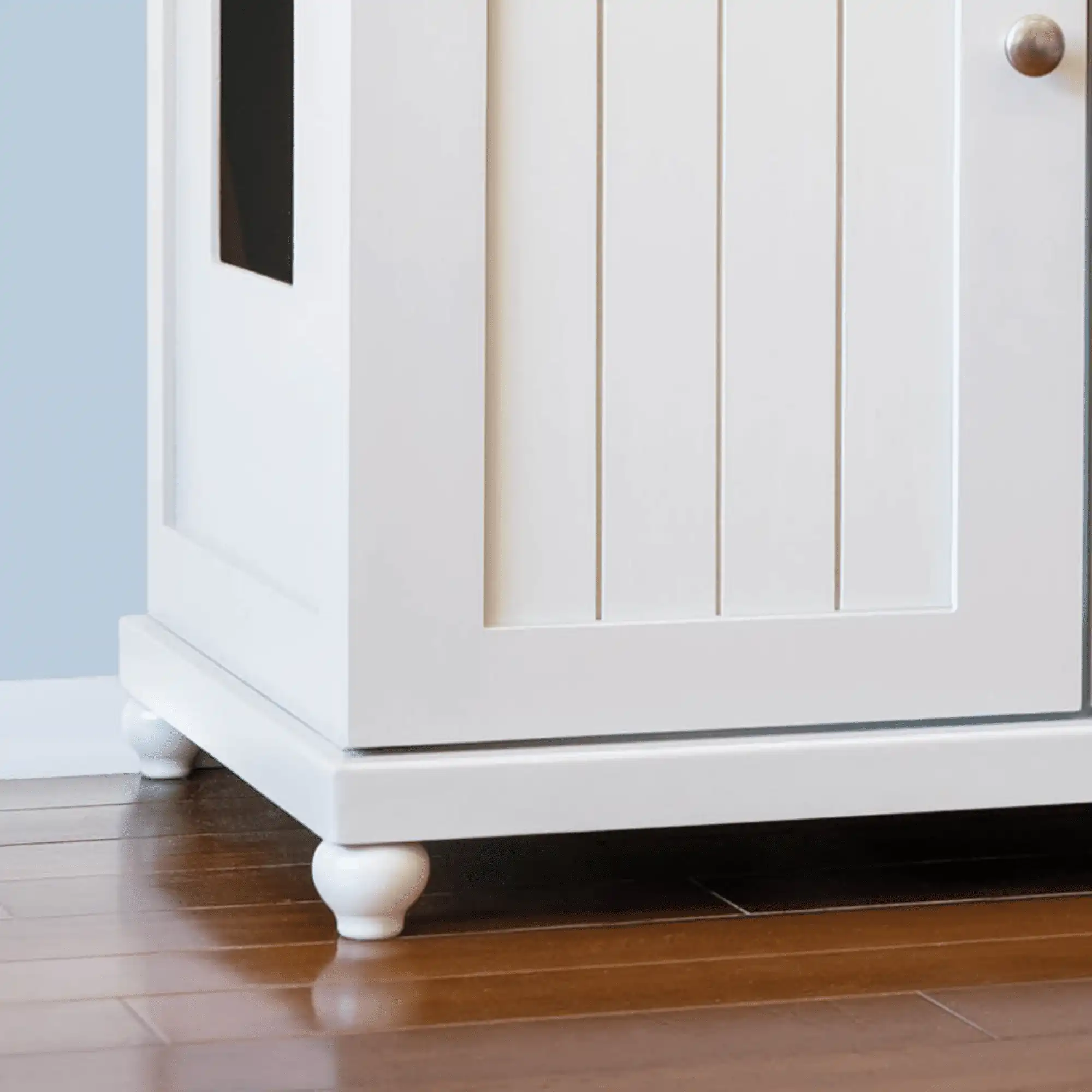 White wooden cabinet with paneled door and round knob, on four tulip-style feet, on wood floor against light blue wall.