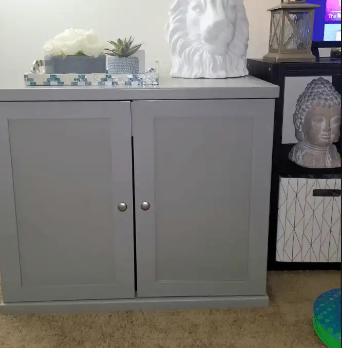 Gray two-door cabinet on carpet with a white lion head sculpture, small plant, and concealed litterbox in a tray.