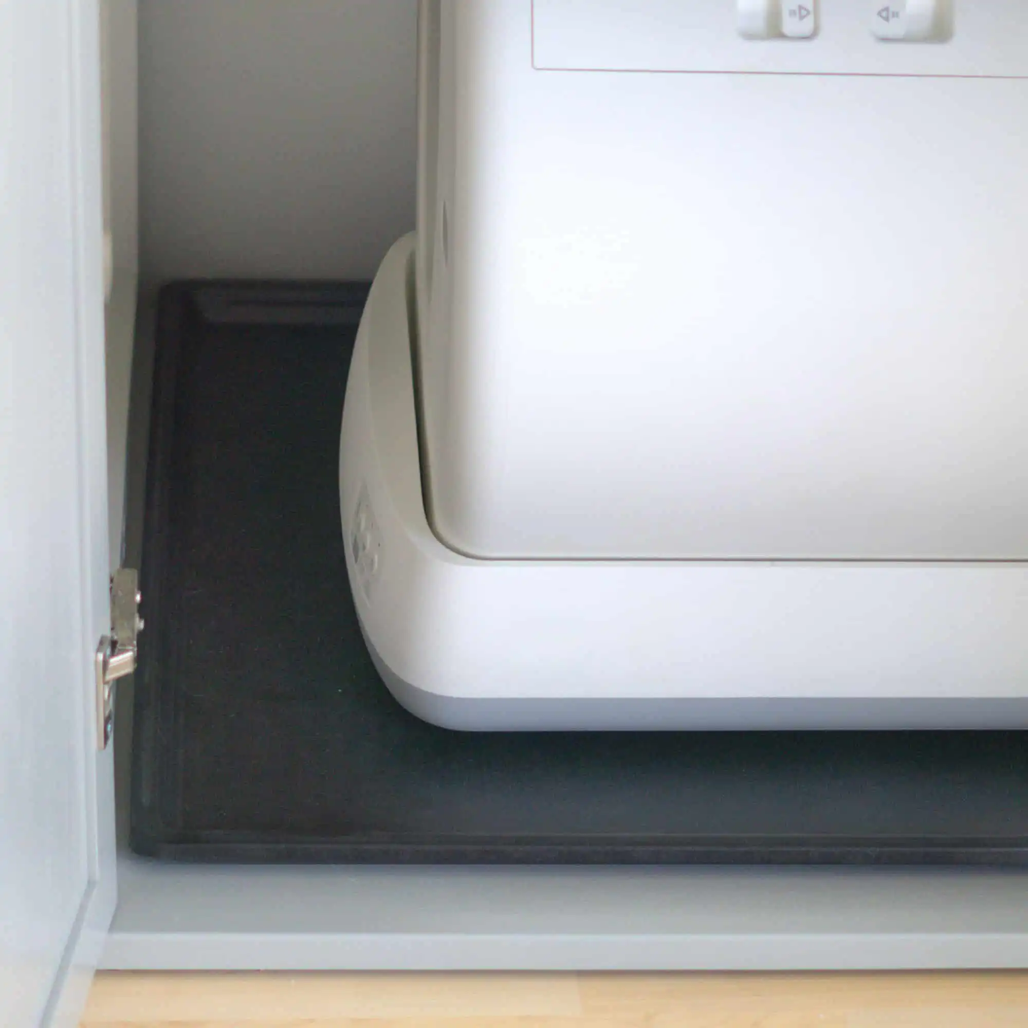 White self-cleaning litter box on a protective tray, placed over a black mat inside an open cabinet, close-up view.