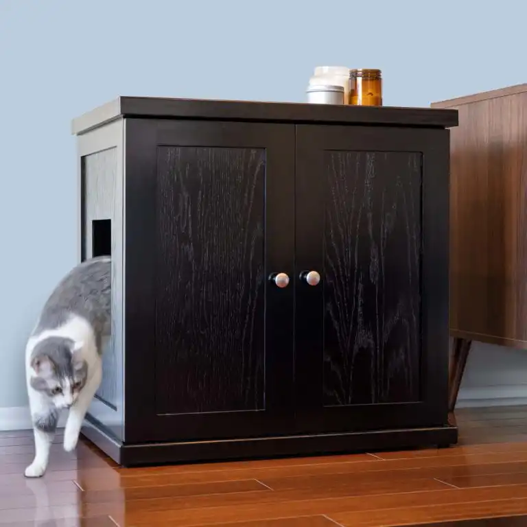 Gray and white cat leaves a black cabinet litter box with side opening on wooden floor beside furniture.