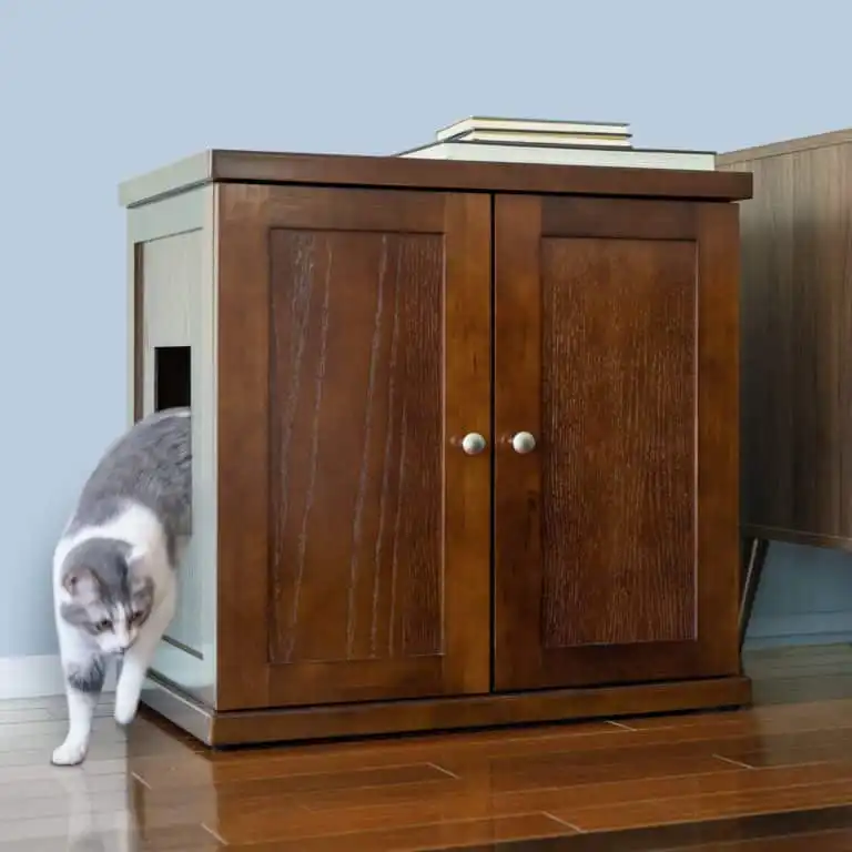 Gray and white cat leaves a wooden litter box cabinet on hardwood floor beside a light blue wall.
