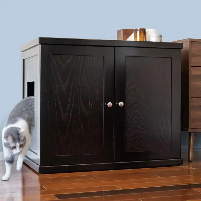 Gray and white cat leaving a litter box cabinet with a side opening on wooden floor beside other furniture.