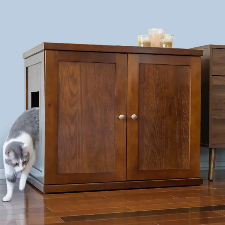 Gray and white cat exits a side door of a wood cabinet with candles on top, on a hardwood floor.