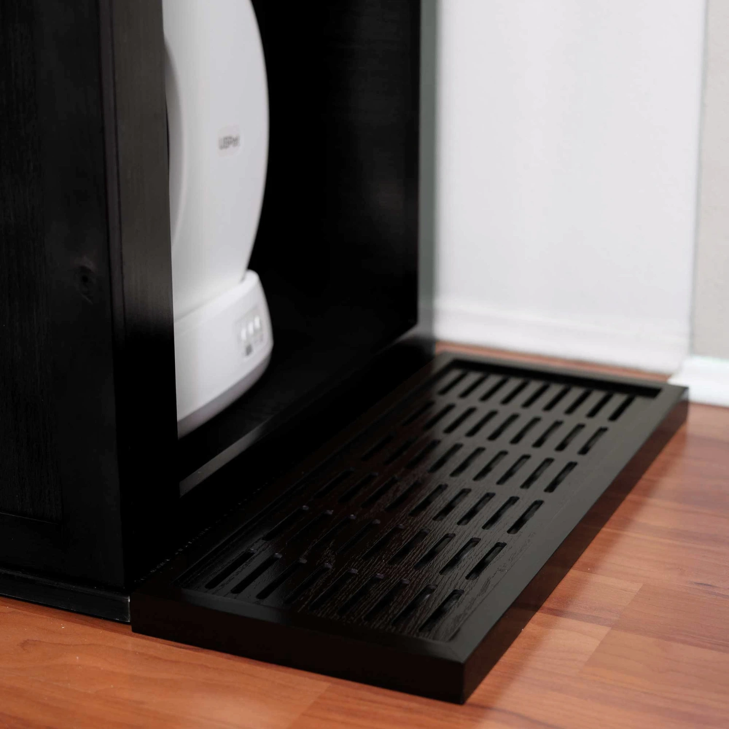 A black slotted plastic mat sits on the floor between a white appliance and a black cabinet, trapping scattered litter.