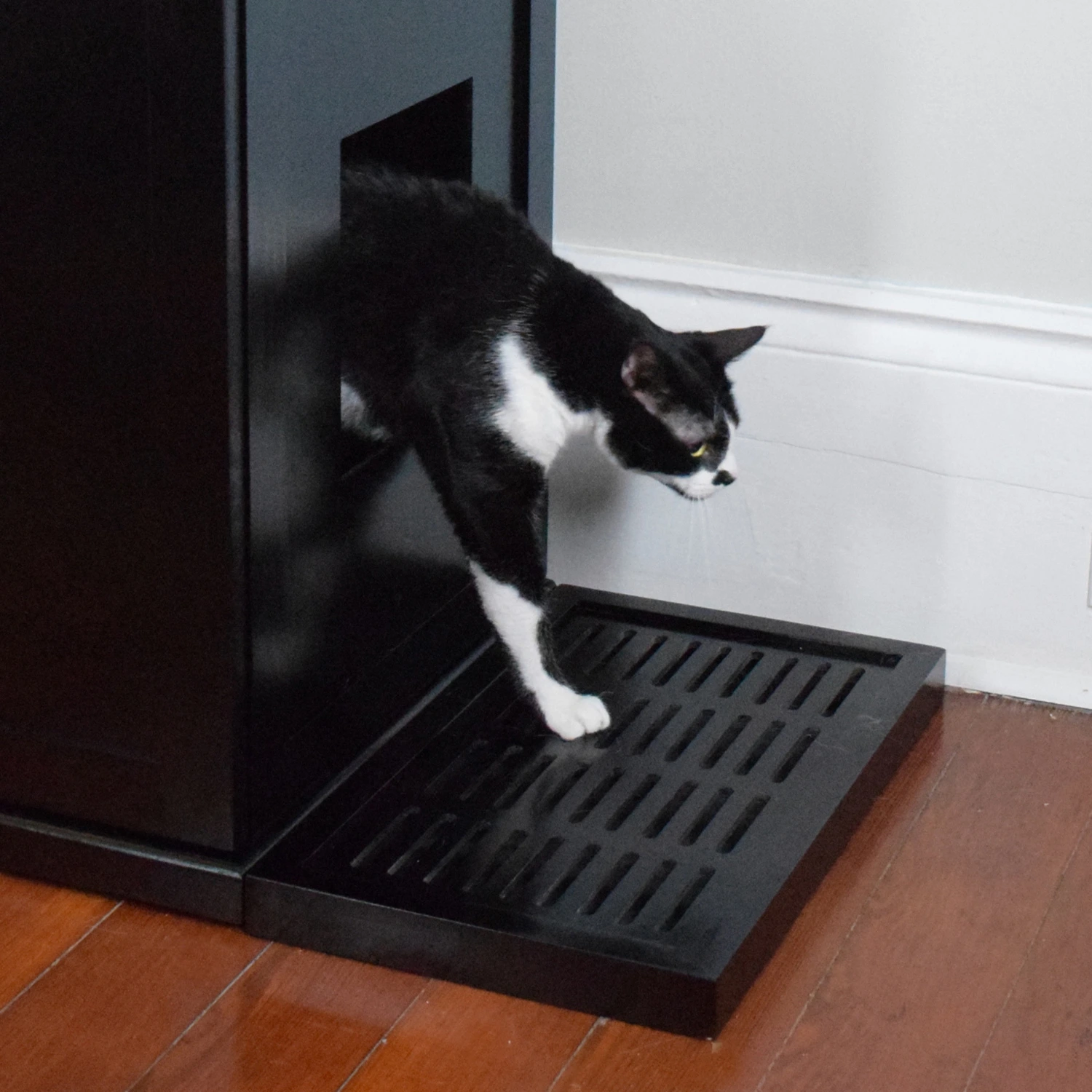 A black and white cat steps from a black litter box cabinet onto an ebony slatted mat on a wooden floor by a white wall.